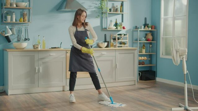 Creative Woman is Washing Floor With Mop Singing and Dancing in Kitchen at Home. Happy Young Woman Cute Housewife is Listening to Music and Dancing in Front of the Fan. Household Chores Concept.