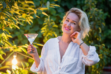 Attractive young blonde woman drinking dry martini in the backyard in the evening. Cocktail...