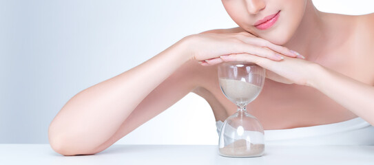 Closeup personable model holding hourglass in beauty concept of anti-aging skincare treatment. Young girl portrait with perfect smooth clean skin and flawless soft makeup in isolated background - obrazy, fototapety, plakaty