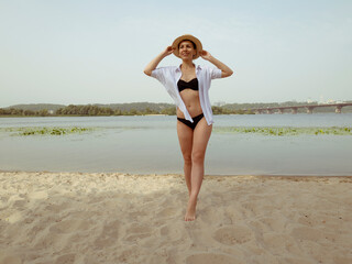 Fototapeta premium A young woman in a dark swimsuit and a hat in a white shirt walks along the beach near the river. Woman smiling while relaxing near the water in summer