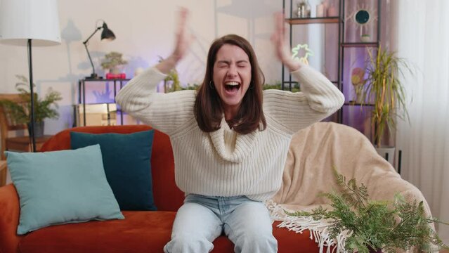 Happy young woman shouting, celebrating success, winning, goal achievement good news raising fists in gesture I did it, lottery luck at modern home apartment. Girl in living room sitting on couch