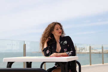 Beautiful blonde woman with curly hair and blue eyes is sitting on the promenade. In the background you can see the sea. The woman looks at camera and makes different expressions, happy, sad.