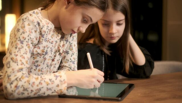 Beautiful teenage girl draws on the touch screen of a digital tablet and talks with her friend at the table, modern tech and generation