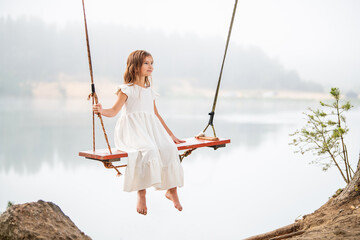 A girl in the forest swinging on a swing. Rope swing on a forest lake. Barefoot girl in a white dress with long hair.