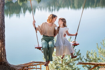 Mom swings with her daughter on a rope swing against the backdrop of a forest lake reflecting the sky. Family concept