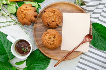 Muffins and cup of herbal tea with blank card, wooden plate, wooden spoon and season herbs on cotton tablecloth.card. Template for recipes or food menu