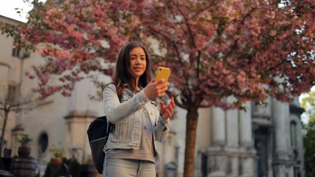 A casually dressed young woman takes a smartphone selfie while standing near a sakura tree with lush spring blossoms. A cute lady is taking pictures for social media while traveling