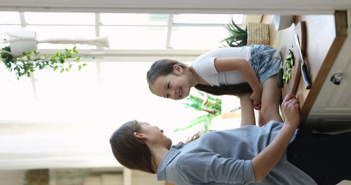 Happy mother cuddling pretty cute girl on kitchen counter, cooking salad for dinner, enjoying motherhood, domestic activity, household work with child, kissing baby girl