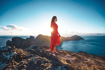 Barefoot woman in red dress enjoys panoramic view over the foothills of a volcanic island and the Ocean. São Lourenço, Madeira Island, Portugal, Europe.
