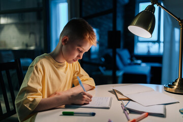 Side view of focused schoolboy studying at home doing homework sitting at table under light of...