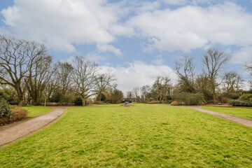 a grassy field with trees in the background and people walking on the path to the right, under a blue sky with white clouds