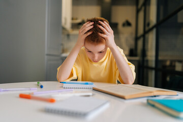 Portrait of exhausted pupil boy tired from studying holding head head with hands sitting at desk...