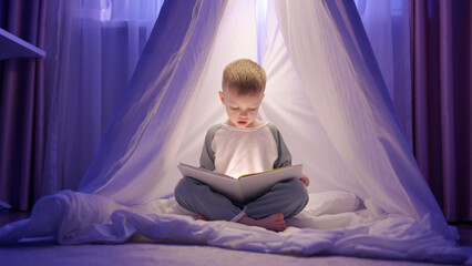 Toddler boy sits in game house and reads aloud glowing book with interest on blanket