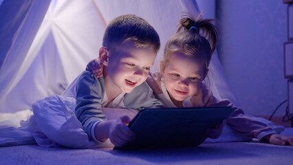 Children lie in game house embracing and laugh watching cartoon on tablet with interest