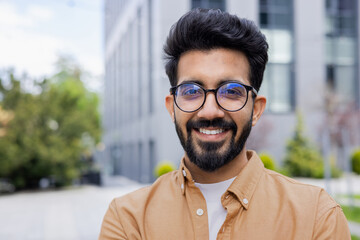 Closeup photo portrait of young entrepreneur wearing glasses, hispanic man smiling and looking at camera, startup entrepreneur outside modern office building.