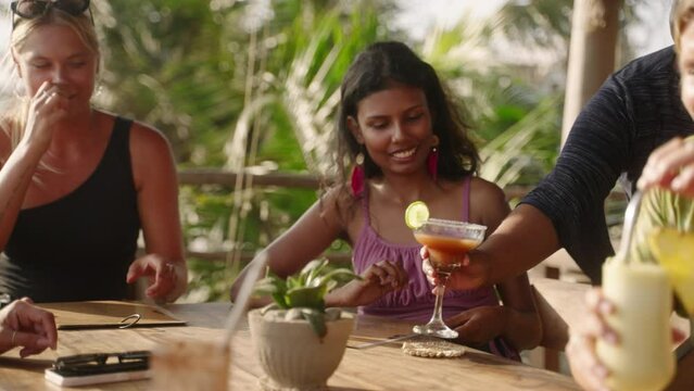 Woman waiter with tray serves cocktails to multiracial women in outdoor tropical restaurant. Female hand takes glasses with alcohol drinks from puts them onto table of multiethnic smiling females
