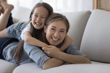 Excited cute Asian girl resting on happy moms back, laughing. Cheerful young mother lying on sofa at home, piggybacking little kid, playing active game with daughter, enjoying motherhood activity