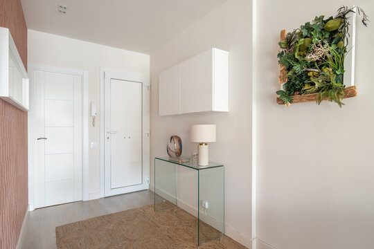 Simple but cozy entrance hall in light colors with an intercom, a glass console and a hanging cabinet with a stylish herbarium on the wall. The concept of simple concise interior design