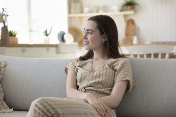 Happy dreamy beautiful young woman sitting on comfortable sofa, relaxing, enjoying comfort, leisure, break, looking away, laughing, smiling at good thoughts, thinking over plans