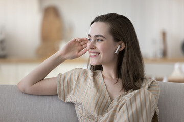 Cheerful young adult girl with small earphones in ears enjoying music on home sofa, looking at window away, smiling, laughing, using wireless audio device, listening to online playlist