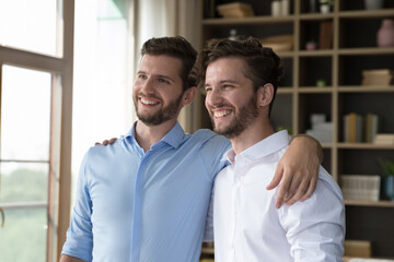 Fototapeta na wymiar Cheerful handsome young adult twin brothers posing for shooting together, standing close, hugging, patting shoulders, looking away, smiling, laughing, enjoying friendship
