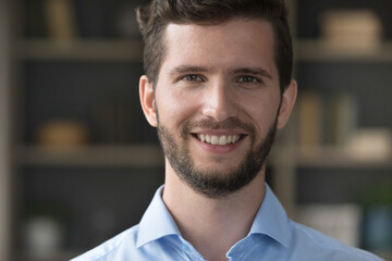 Positive attractive young man close up facial portrait. Face of happy handsome guy with trendy stubble posing indoors, looking at camera with toothy smile. Cropped shot