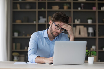 Tired sad freelancer man getting problems with laptop, software error, feeling stress, exhaustion, headache, touching head at home work table, looking at screen with doubt