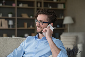 Positive handsome young freelancer, business man in glasses talking on cellphone at home, making telephone call, looking away, listening, speaking, smiling, laughing