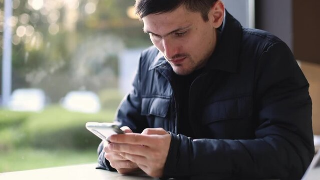 One young handsome Caucasian brunette man in a dark jacket sits at a table by the window in a diner and carefully looks at the phone screen in his hands, side view close-up.