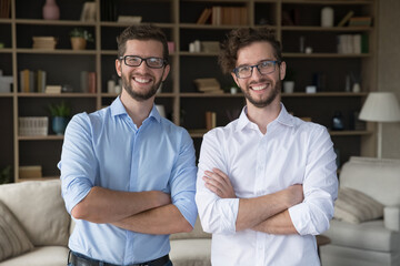 Positive confident family business partner men portrait. Happy handsome young adult twin brothers in glasses and office shirts posing together at home with arms crossed, looking at camera, smiling - obrazy, fototapety, plakaty