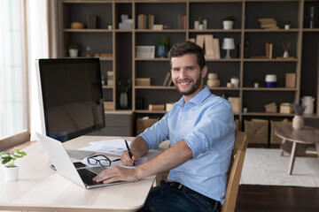 Happy freelance professional programmer sitting at table with workstation computer, typing on laptop, posing at home work table, looking at camera, smiling. Young freelancer portrait