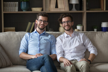 Happy positive young adult identical twin men in glasses sitting together on couch, looking at camera, smiling, meeting at home for family event, keeping good close relationships. Indoor portrait