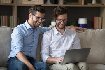 Cheerful young twin brother men in glasses using laptop for Internet job communication, working at home together, brainstorming on freelance business project