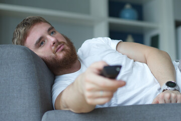 portrait of boring young man in casual clothes sitting and lay on sofa in living room and looking tv with remote control in hand at home 