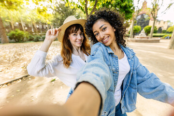 Fototapeta na wymiar Two young trendy multiracial women having fun riding a bike together on summer vacation. Female friends taking selfie portrait together enjoying day outside. Friendship and youth lifestyle concept.