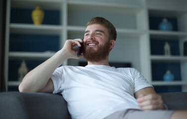 Happy young man talk, chat on cell mobile phone, smile, have fun, sit on comfortable couch at home in living room, call on smartphone, relax 