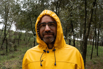 portrait of a man with glasses and beard wearing a bright yellow hooded raincoat in the middle of the forest.