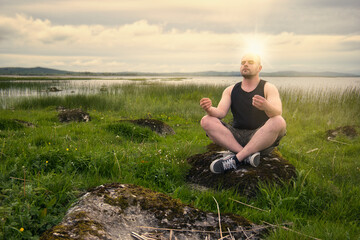 Caucasian man in sleeveless shirt and camo shorts meditating on the rock in the field by the Corrib lake in Galway, Ireland 
