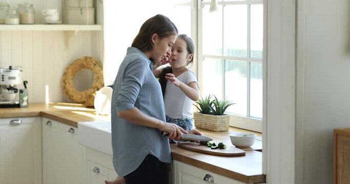 Caring mom and sweet little daughter girl cooking salad in kitchen, chopping fresh vegetables on counter at window, eating pieces, talking, kissing, enjoying family leisure culinary hobby