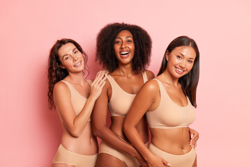Three very happy girlfriends wearing beige tops and panties pose inside, black-skined girl in the middle her friends are on both sides, comfort underwear concept, copy space - obrazy, fototapety, plakaty