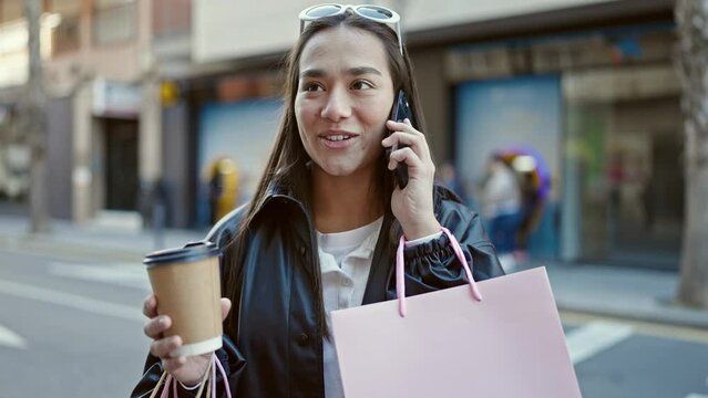 Young beautiful hispanic woman talking on smartphone holding shopping bags and coffee at street