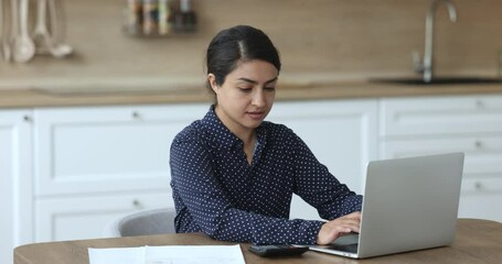 Focused young Indian woman sit in kitchen make remotely work with calculator and laptop calculate income, expenses, estimate family budget, accountant conduct financial inspection, summarize total sum - Powered by Adobe