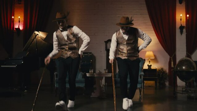 Two stylish professional male dancers in dandy hat walking to camera in music beat and throwing each other classic cane, jumping synchronically performing jazz funk dance moves for night show event 4K
