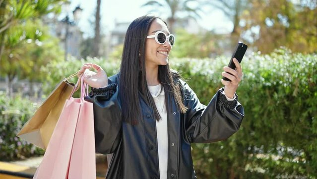Young beautiful hispanic woman make selfie by smartphone holding shopping bags at park