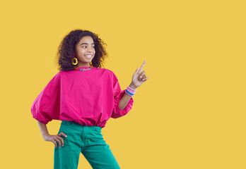 Advertise here. Beautiful and stylish ethnic preteen girl shows copy space for advertisement on yellow background. Cute smiling curly dark-skinned teen girl advertising or recommending product. Banner
