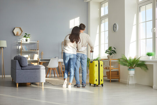 View from behind of hugging loving couple standing with suitcase at home. Full length shot of couple wearing casual clothes leaving their home with luggage to go on vacation