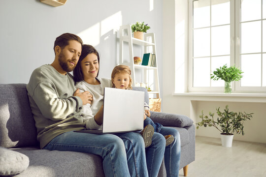 Happy family sitting on couch using laptop at home. Mom, dad and cute toddler baby having video call, watching movie or shopping online at home. Family having good time at home