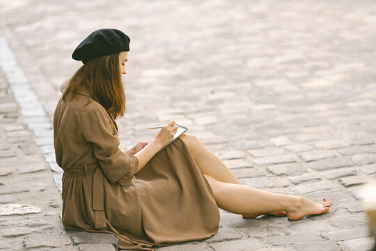 Girl in dress and black hat painting a sketch while sitting on a street
