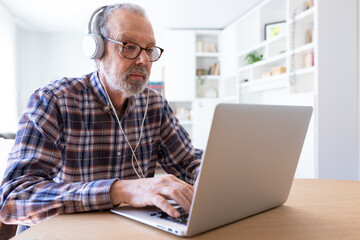 Senior man working with laptop and using headphones at home dining room.