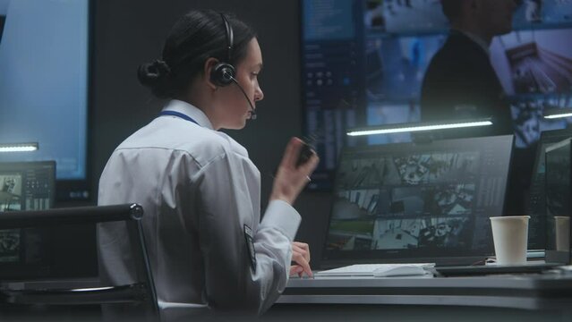 Female security officer in headset works on computer with CCTV cameras video footage in police monitoring center, talks in radio set. Male employee controls surveillance cameras on big digital screen.